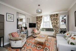 Ein Wohnzimmer mit Sofa, Sesseln, Hocker, Fernseher und einem gemusterten Teppich.