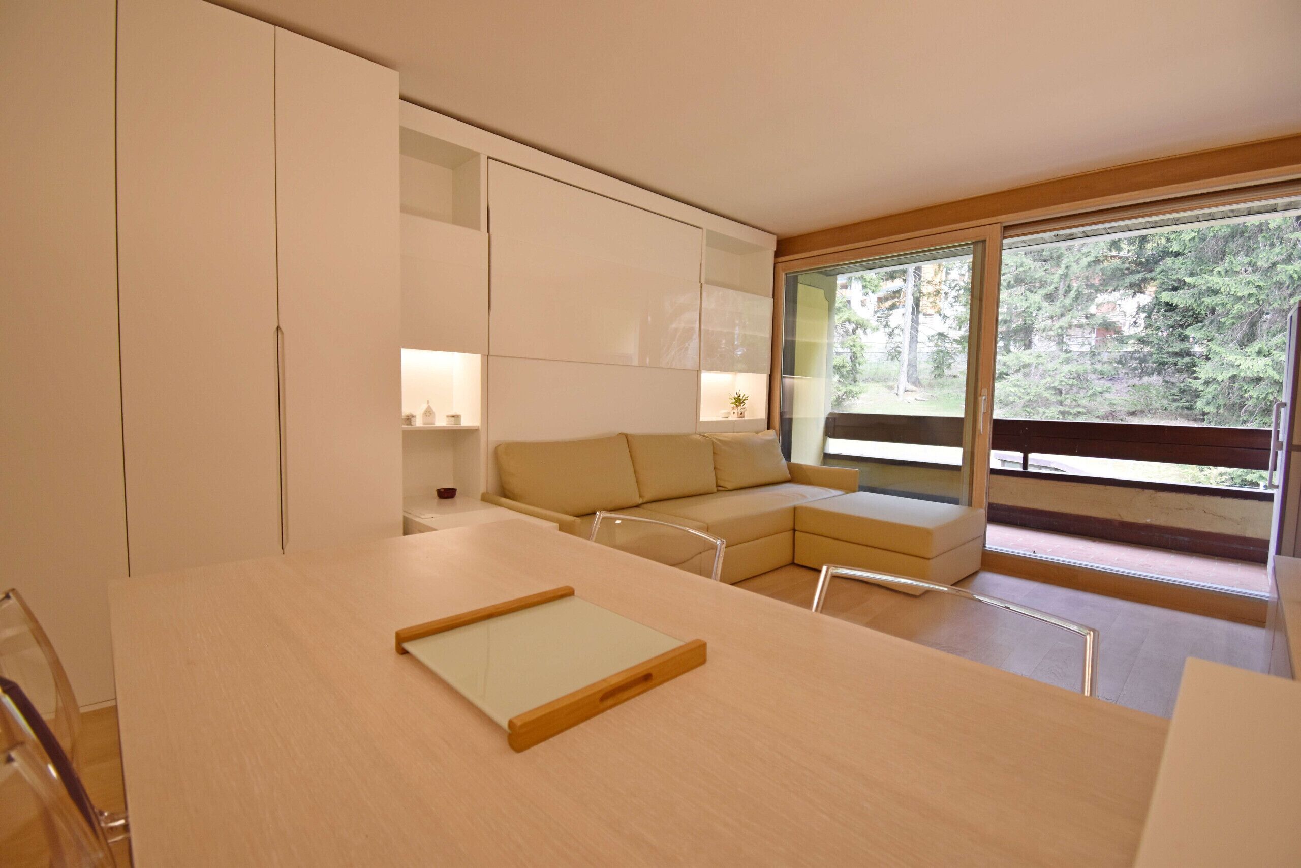 A modern interior with a wooden table, a beige sofa, and a large window with a view of greenery.