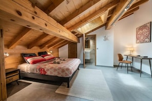 Ein Schlafzimmer mit Holzboden, einem Bett mit Blumenmuster-Bettwäsche, einem Schreibtisch mit Stuhl und einem kleinen Badezimmer, das durch eine offene Tür sichtbar ist.