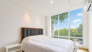 Een slaapkamer met een bed, een nachtkastje, een raam met uitzicht op bomen en gebouwen, en inbouwspots.