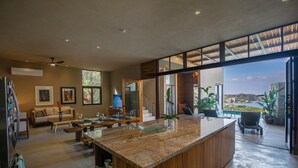 Une cuisine spacieuse avec un grand îlot central, un coin repas avec un canapé et une vue sur un balcon agrémenté de plantes et d’une piscine.