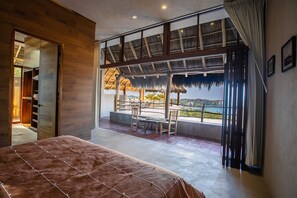 Ein Schlafzimmer mit einem großen Bett, Blick auf einen Balkon mit Tisch und Stühlen sowie einem Strohdachhaus.