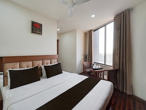 A hotel room with a large bed, a chair, a table, a window with curtains, a ceiling fan, and a painting on the wall. - Super Deluxe Roomhotel Navjivan (Himatnagar)