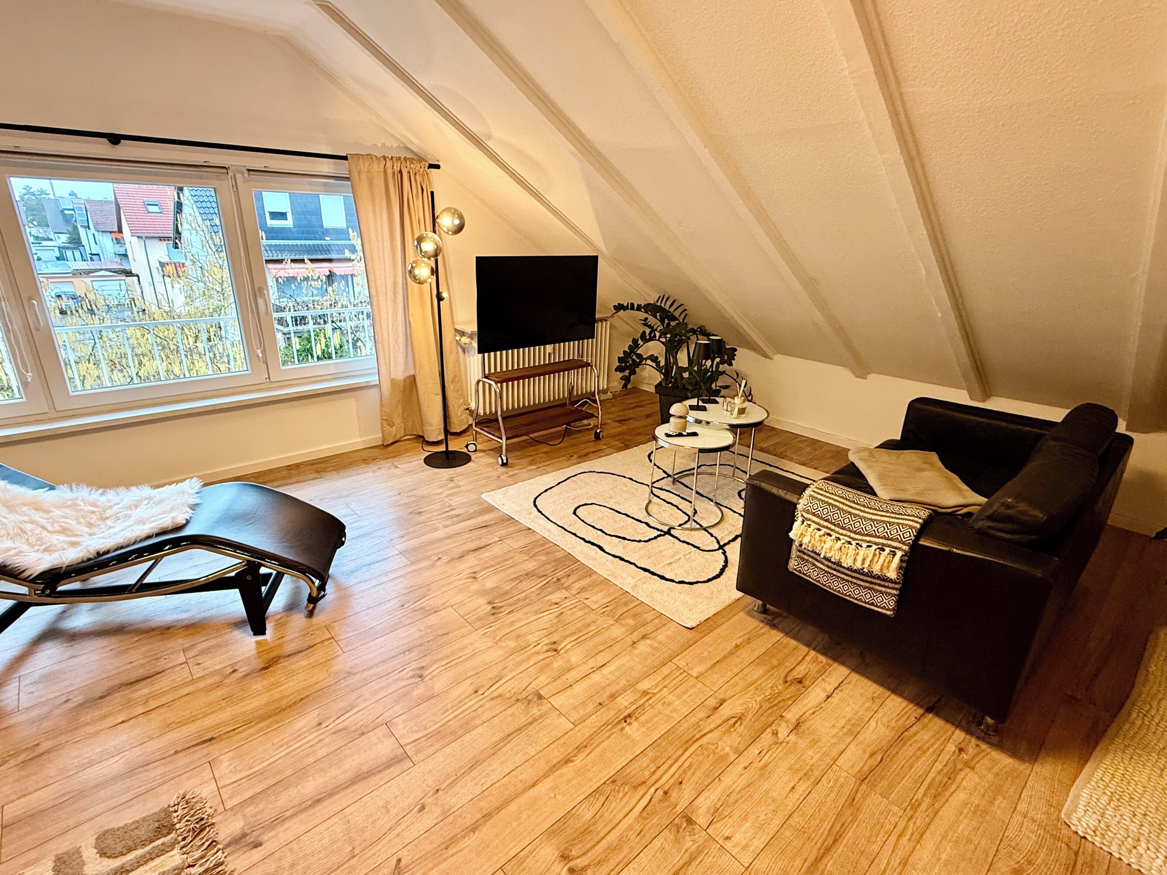 Un appartement moderne de style « loft », doté d’une grande fenêtre, d’un canapé en cuir noir, d’un tapis blanc et d’un lampadaire noir.