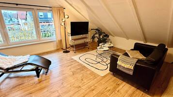 Un appartement moderne de style « loft », doté d’une grande fenêtre, d’un canapé en cuir noir, d’un tapis blanc et d’un lampadaire noir.