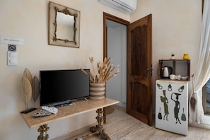 A room with a television on a wooden table, a mirror on the wall, an air conditioner, a door, and a refrigerator with decorative figures.