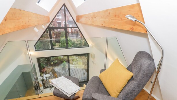 A modern loft bedroom with a sloped ceiling, a large skylight, a grey armchair with a yellow pillow, a wooden floor, and a glass partition. - Little Mornington (Malvern)