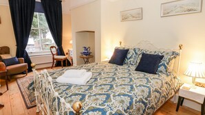 A bedroom with a patterned bedspread, a white radiator, a chair, a table with a pitcher, and a window with curtains. - Little Mornington (Malvern)