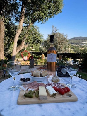 A table with a bottle of wine, various cheeses, meats, and pickles, set outdoors with trees and a view of a hill in the background.