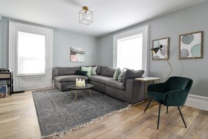 A living room with a gray sectional sofa, a round coffee table, a dark green chair, a patterned rug, and artwork on the walls.
