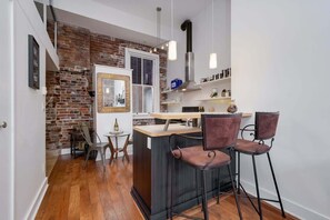 Une cuisine compacte avec un mur en briques, un petit coin repas avec des tabourets de bar et une fenêtre avec des stores.