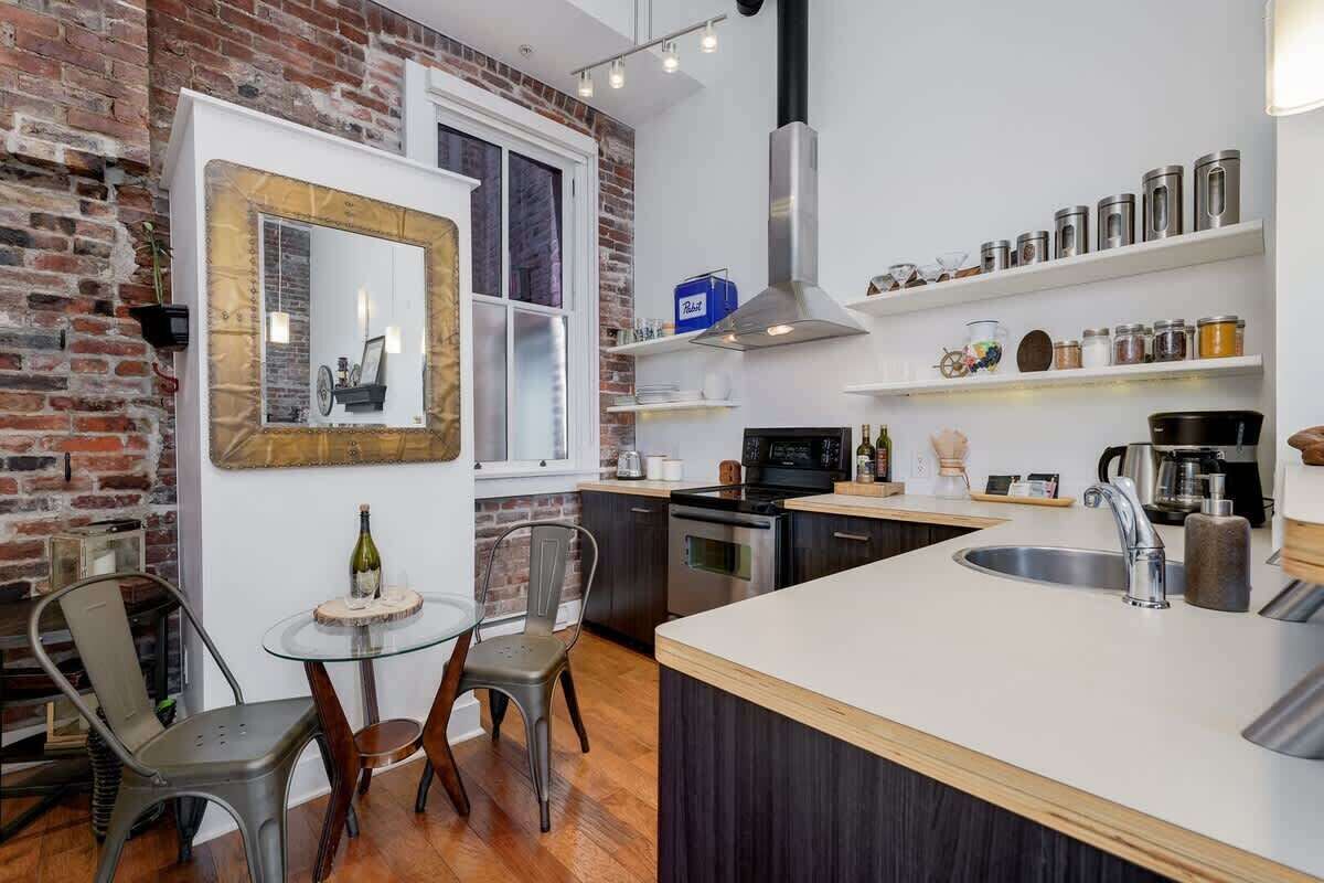 Een keuken met een bakstenen muur, een grote spiegel, een ronde eettafel met stoelen, een centraal keukeneiland met een spoelbak en roestvrijstalen apparatuur.