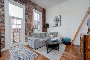 Un salon avec un canapé gris, une table basse, un tapis à motifs, un meuble en bois et une grande fenêtre donnant sur un immeuble en briques.