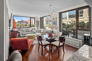 A spacious living area with a dining table, comfortable seating, a chandelier, and large windows offering views of the cityscape.