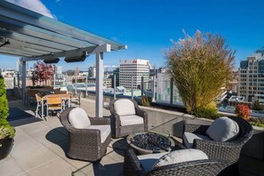A rooftop terrace with wicker furniture, a glass table, and a view of city buildings.
