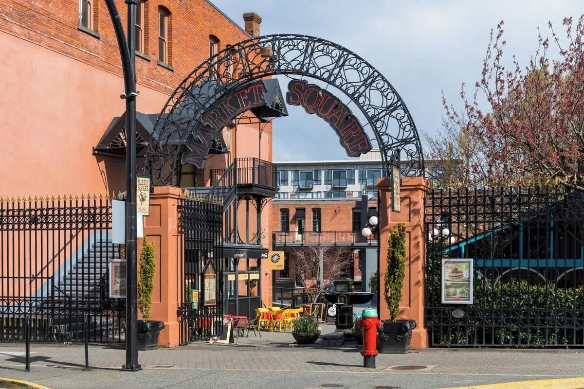 Um portão de ferro forjado com a inscrição 'Market Square' acima, que dá acesso a um edifício com fachada de tijolos e uma escadaria.