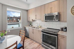 A modern kitchen with stainless steel appliances, wooden cabinets, a white tiled backsplash, and a window with a view of buildings. - Cozy Downtown Victoria Condo (Victoria)