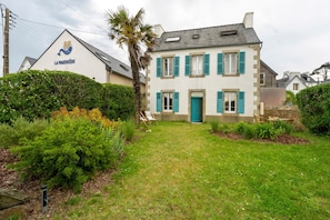 Un bâtiment à deux étages, doté d’une façade blanche et de volets bleus, avec un palmier et une pelouse bien entretenue.