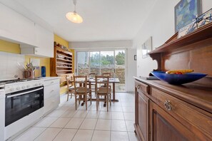 Une cuisine avec des meubles blancs, une cuisinière, une table à manger avec des chaises et une grande fenêtre.