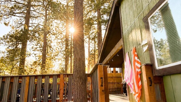 A hot tub in a forested area with a wooden deck and a building with large windows. - 3bdr Modern Family House - Spa, Playroom, Walkable, Pet-friendly (Big Bear Lake)