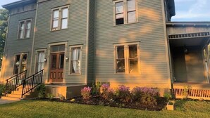 Uma casa de dois andares com varanda da frente, escada e um pequeno jardim com flores.