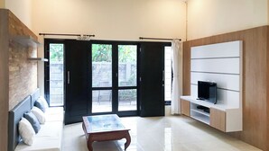 Ein modernes Wohnzimmer mit Sofa, Couchtisch, Fernseher auf einer Holzablage und Glastür zum Garten.