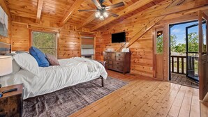 A wooden cabin bedroom with a bed, dresser, TV, and a balcony with a view. - Douglas Lake Log Cabin | Game Room, Big Yard, Sleeps 6 (Sevierville)