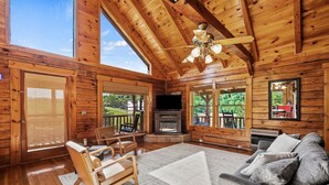 A log cabin interior with a fireplace, a sofa, a chair, and large windows. - Douglas Lake Log Cabin | Game Room, Big Yard, Sleeps 6 (Sevierville)