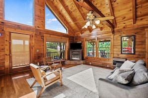 A log cabin interior with a fireplace, a sofa, a chair, and large windows. - Refreshed 2BR Cabin w/ Game Room | Big Yard | Douglas Lake Area, Sevierville (Sevierville)