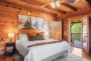 A wooden cabin bedroom with a bed, nightstand, lamp, and a painting of a mountain landscape. - Refreshed 2BR Cabin w/ Game Room | Big Yard | Douglas Lake Area, Sevierville (Sevierville)