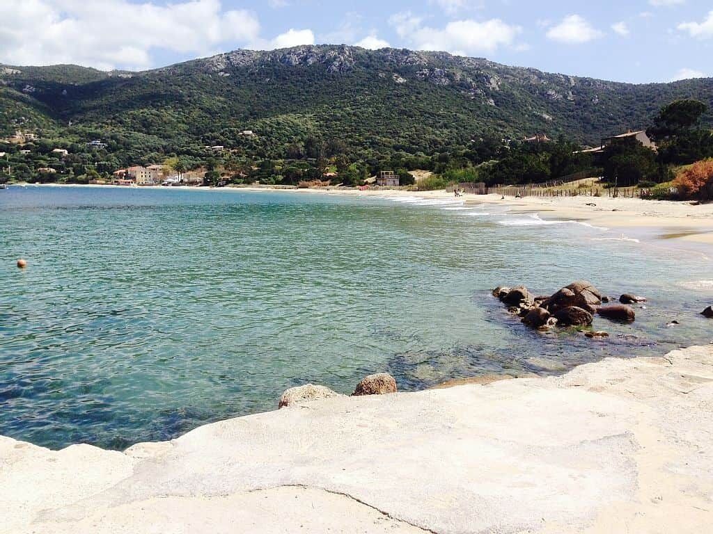 Une plage à l’eau claire, avec un rivage rocheux et une chaîne de montagnes en arrière-plan.