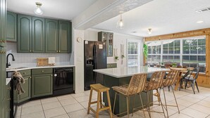 A kitchen with green cabinets, a black refrigerator, a wooden island with bar stools, and a window with blinds. - Aloha Pool House- Stay Local, Live Aloha (Fort Walton Beach)