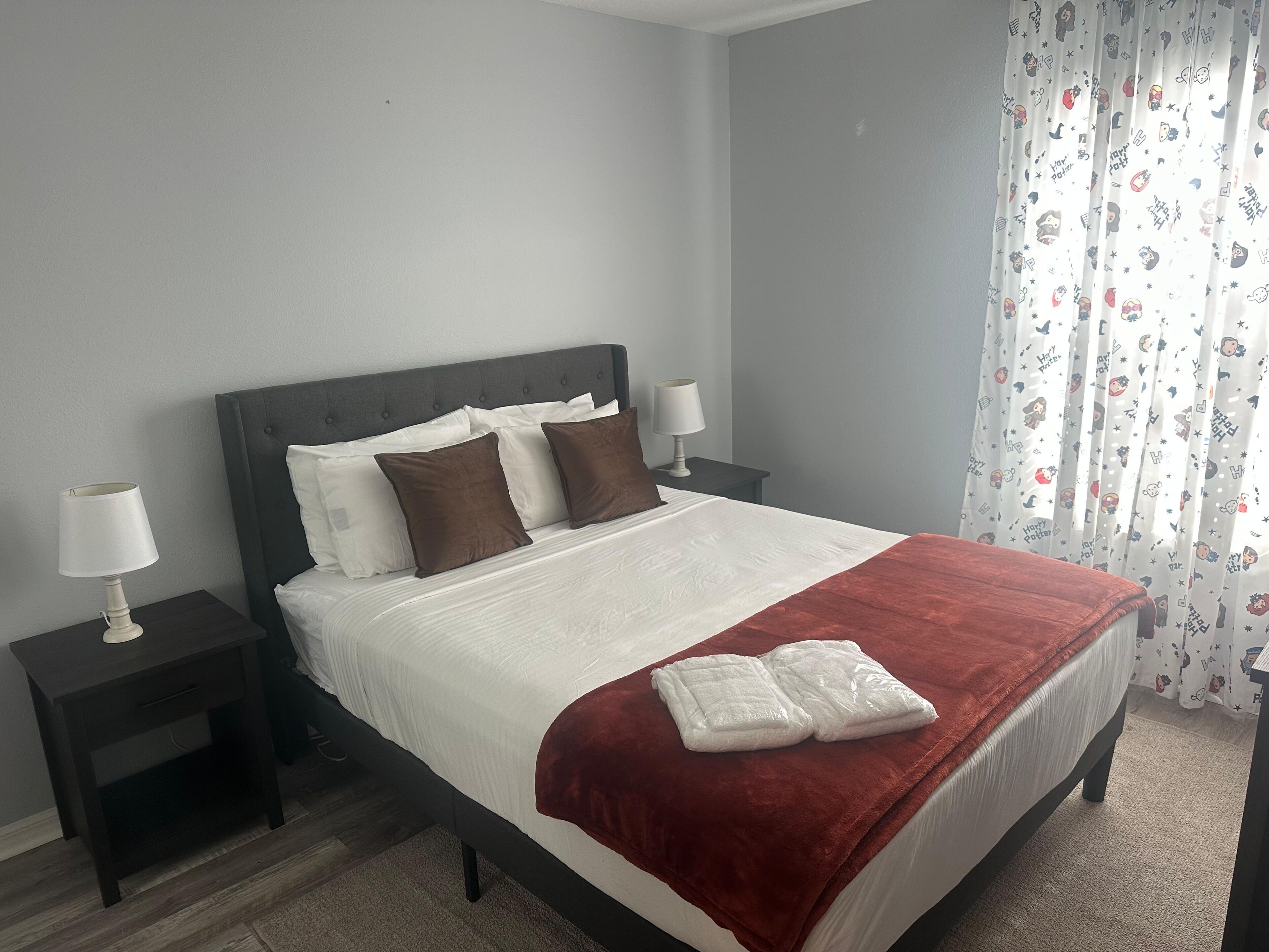 A neatly made bed with a red blanket, two bedside tables with lamps, and a window with curtains.