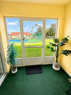 A room with a sliding glass door leading to a tennis court, two potted plants, and a welcome mat.