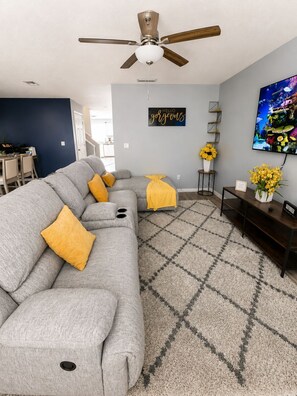 A living room with a sectional sofa, a ceiling fan, a wall-mounted TV, and a decorative sign.