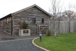 A rustic wooden building with a sloped roof, a gated entrance, and a small planter with a sign. - Rushmoor Retreat, Cosy with Outdoor Pizza Oven (Bratton)