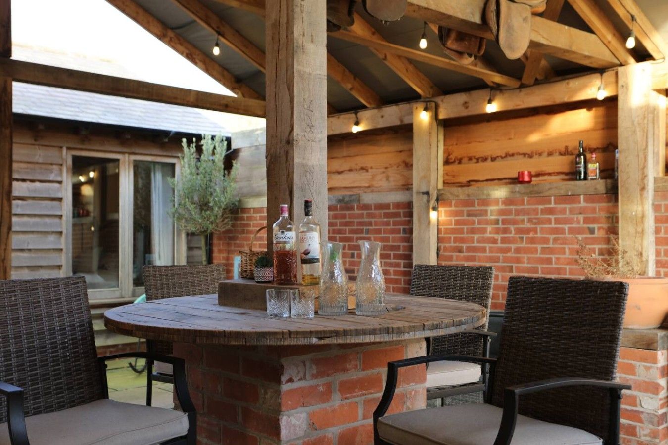 Une terrasse extérieure avec une table ronde en bois, des chaises en osier et un mur en briques.