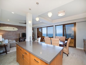 A spacious living area with a large island, a dining table with wicker chairs, a surfboard on the wall, and a view of the sea through large windows. - Makena Surf F207 Beachfront Resort (Wailea)