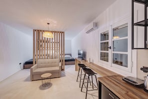 Un espacio habitable moderno con un sofá, una zona de comedor con taburetes, un televisor y una cocina con hervidor.