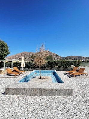 A swimming pool with lounge chairs and umbrellas, surrounded by pebbles and a mountain in the background.