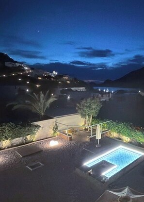 Vista nocturna de una zona costera con piscina, mobiliario exterior y edificios a lo lejos.