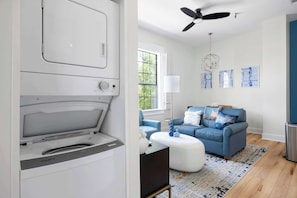 A modern living room with a large window, a ceiling fan, a blue sofa, and a white washing machine. - 2BR Retro Gaming Retreat | Near Stadium (Jacksonville)