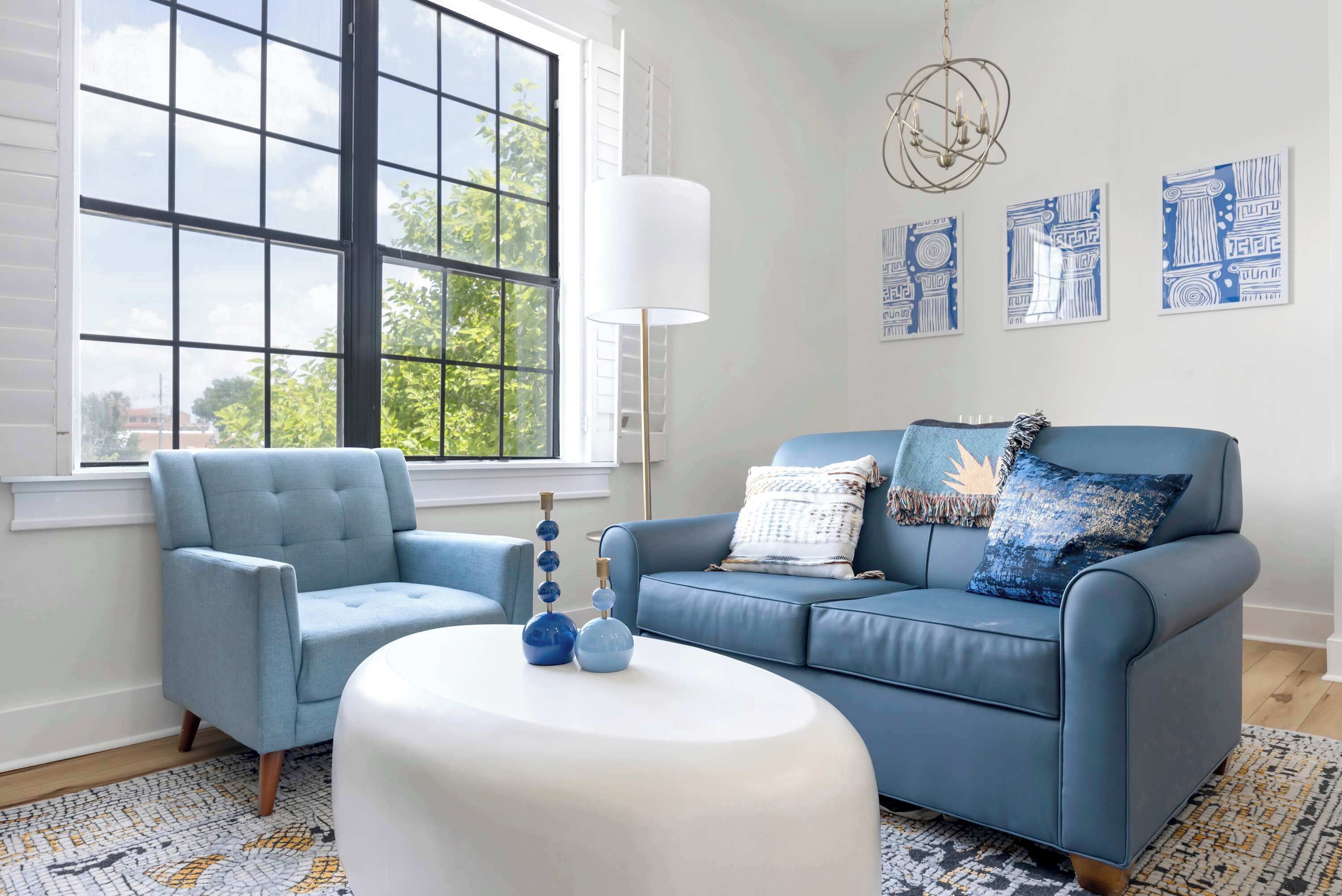 A living room with a blue sofa, a blue armchair, a white ottoman, and a large window with a view of trees.