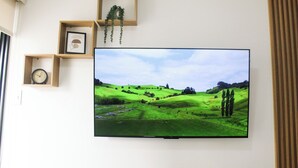 A flat-screen TV mounted on a wall with a landscape image, wooden shelves with a clock and framed picture, and a window with blinds. - Stylish Studio in Bohemian Barranco · Work-Ready · Steps from Miraflores (Barranco)