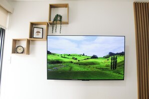 Un televisor de pantalla plana montado en la pared, con una imagen de paisaje, estantes de madera con un reloj y un cuadro enmarcado, y una ventana con persianas.