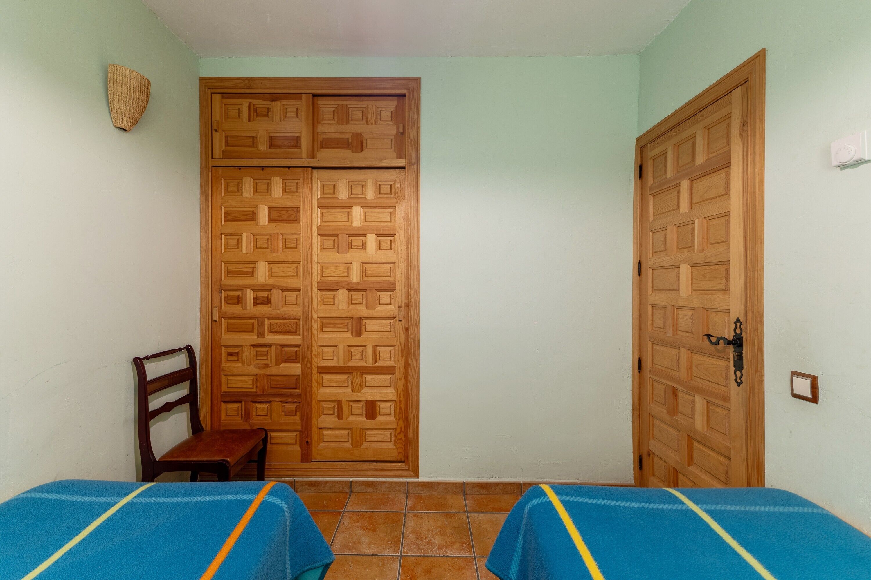 Une chambre avec deux lits, une armoire en bois, une chaise et un luminaire fixé au mur.
