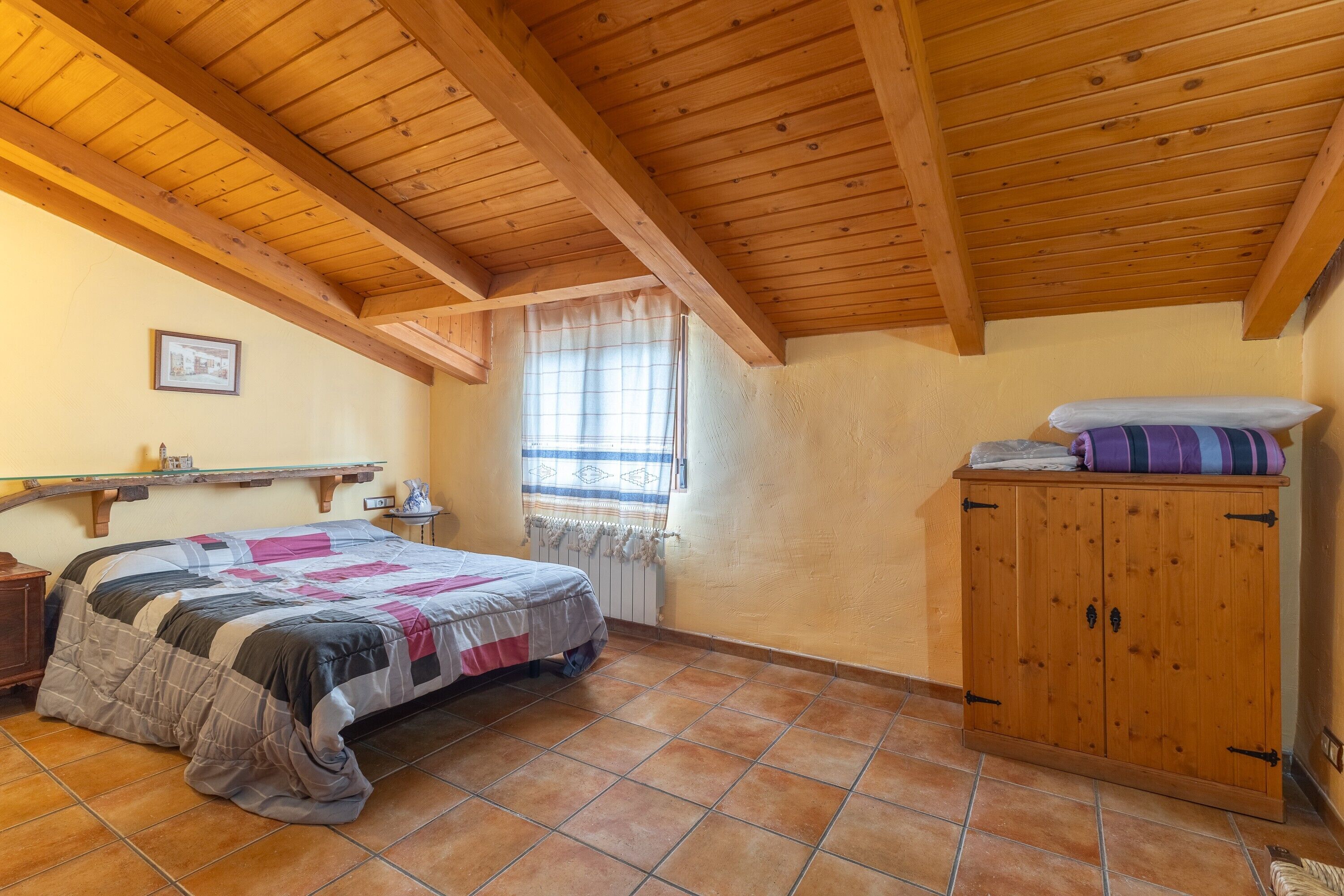 Une chambre à coucher avec un lit, des poutres en bois au plafond, une fenêtre avec des rideaux, une armoire en bois et un sol carrelé.