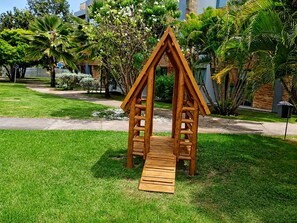 A wooden playhouse with a ramp in a well-maintained garden area. -  Iloa Resort Espaço para Famílias e Grupos; 3 Q e 2 Vagas (Barra de São Miguel)