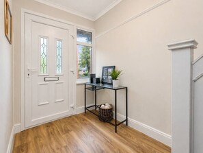 A hallway with a white door, a window with a view, a black metal console table with a plant, and a basket with decorative items. - Wembley Garden Retreat |4BR|Sleeps 7|Near Station (Greater London)