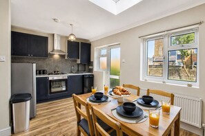 A modern kitchen with dark cabinets, a stainless steel refrigerator, and a dining area with a wooden table set for breakfast. - Wembley Garden Retreat |4BR|Sleeps 7|Near Station (Greater London)